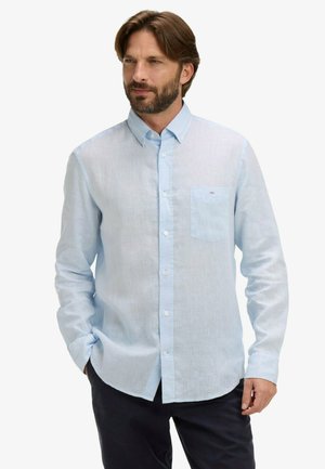 Homme barbu portant une chemise à manches longues bleu clair boutonnée et un pantalon foncé, debout les mains dans les poches, regardant sur le côté.
