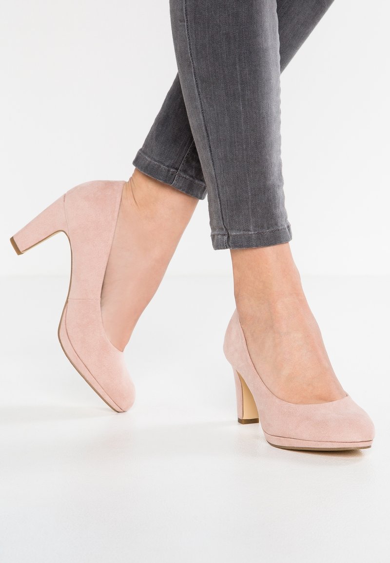 Femme portant des escarpins à talons hauts en daim rose clair associés à un jean skinny gris foncé retroussé, sur fond blanc.