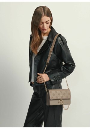 Kobieta o długich brązowych włosach, ubrana w czarną skórzaną kurtkę, czarne spodnie, białą koszulę oraz beżową torebkę na łańcuszku.