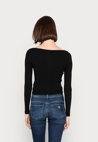 Top nero a maniche lunghe con coste e scollo a barca, abbinato a jeans blu aderenti con dettagli sulle tasche posteriori e una patch con logo triangolare.