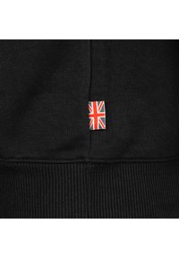 Melna auduma ar ribotu apmali un nelielu etiķeti, kurā izšūta sarkanbalti zila Lielbritānijas karoga Union Jack motīva piešuve pie šuves.