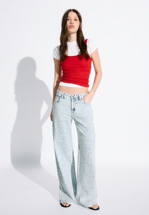 Young woman with long brown hair wearing a red tank top over a white T-shirt and loose light blue jeans, standing with one hand in pocket.