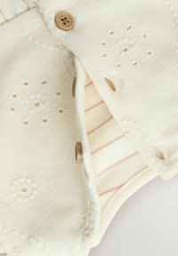 Vêtement en coton blanc avec des détails brodés, présentant des boutons en bois et une doublure intérieure rayée. Texture douce, forme arrondie.