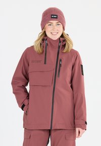 Bordeauxfarbene, wasserdichte Jacke mit Reißverschluss an der Vorderseite, großer Schultertasche und verstellbarer Kapuze. Begleitet von einer passenden Strickmütze.