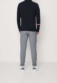 Maglione girocollo blu navy con colletto alto, caratterizzato da una striscia rossa e bianca sulla manica. Abbinato a pantaloni a quadri grigi chiari e sneakers bianche.