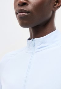 Κοντινό πλάνο ενός ατόμου με σκούρο δέρμα που φοράει ένα ανοιχτό μπλε μπλουζάκι με φερμουάρ και μικρά χρυσά σκουλαρίκια σε σχήμα κρίκου, με φόντο απλό.