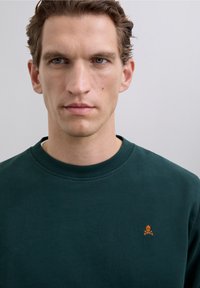 Bărbat cu păr scurt și brunet, purtând un pulover de culoare verde închis, cu guler rotund, care are un logo mic cu un craniu și oase încrucișate portocalii pe piept.