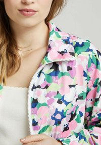Veste légère à motif floral coloré avec fermeture éclair. Présente des accents roses, verts, noirs et bleus sur un fond blanc.