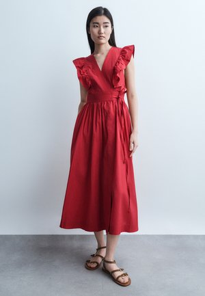 Γυναίκα που στέκεται, φοράει αμάνικο κόκκινο φόρεμα τύπου wrap με βολάν στους ώμους και καφέ επίπεδα σανδάλια, πάνω σε απλό γκρι φόντο.