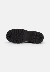 Suela de bota de goma negra con surcos profundos y patrones de tracción variados. Incluye un detalle de logo rectángulo cerca del talón.