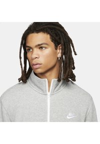 Γκρι φούτερ με φερμουάρ και ψηλό γιακά, κατασκευασμένο από μαλακό ύφασμα; διαθέτει το λογότυπο της Nike σε λευκό χρώμα στο αριστερό στήθος και λευκό φερμουάρ.