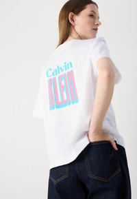 T-shirt en coton blanc à coupe décontractée, avec un texte audacieux en rose et turquoise indiquant "Calvin KLEIN" dans le dos. Pantalon en denim foncé.