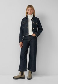 Veste en denim foncé avec des boutons dorés et deux poches avant, portée avec un col roulé blanc et un pantalon en denim à jambe large. Bottines grises.