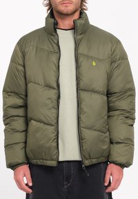 Olivengroen pufferjack met hoge kraag, horizontaal gestikte textuur, frontrits en een klein logo-accent op de borst.