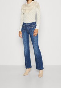 Hellbeiger gestreifter Strickpullover mit geometrischen Mustern und langen Ärmeln, kombiniert mit blauen ausgestellten Jeans mit goldfarbenen Akzenttaschen.