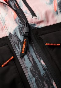 Chaqueta colorida con un patrón abstracto en rosa y negro, secciones de tela negra y cremalleras y detalles naranjas para mayor visibilidad.