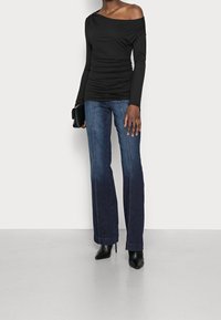 Sort off-shoulder top med samlet tekstur, parret med mørkeblå wide-leg jeans, der har synlige sømme, og sorte spidse hæle.