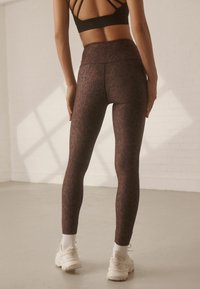 Hoch taillierte Leggings mit einem dunklen lila Muster, aus dehnbarem Stoff. Kombiniert mit weißen Turnschuhen und Knöchelsocken.