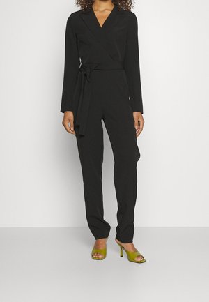 Woman wearing a black long-sleeve wrap jumpsuit with a tied waist and green open-toe heeled mules, standing against a plain white background.