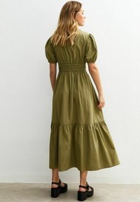 Vestido largo de color verde oliva con mangas abullonadas, cintura fruncida y dobladillo en capas. Combinado con sandalias de plataforma negras. Textura suave, material ligero.