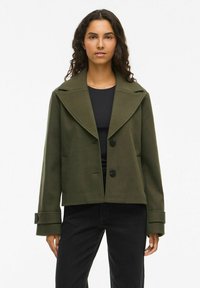 Olijfgroen cropped jack met een brede revers, twee zwarte knopen en zijzakken. Gelaagde stof, gecombineerd met een zwarte top en jeans.