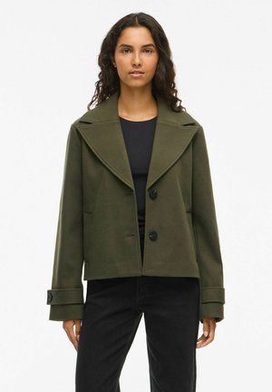 Veste courte olive avec un large col, deux boutons noirs et des poches avant. Tissu texturé, associée à un haut noir et à un jean.