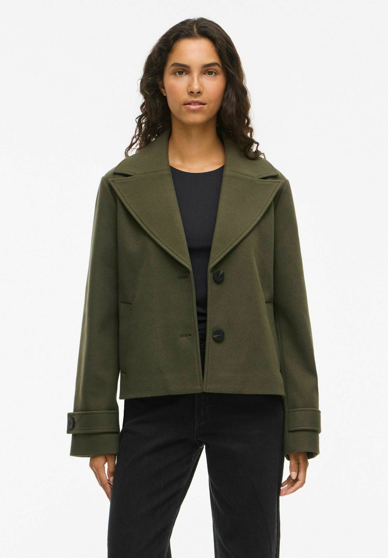 Olijfgroen cropped jack met een brede revers, twee zwarte knopen en zijzakken. Gelaagde stof, gecombineerd met een zwarte top en jeans.