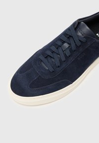Sapatilha de camurça azul-marinho com atacadores texturizados e costura tonal. Apresenta uma sola de borracha em tom de creme e logótipo em relevo no calcanhar.