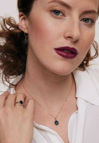 Złoty naszyjnik z niebieskim kwiatowym wisiorem, w komplecie z pasującymi niebieskimi kolczykami. Modelka nosi także pierścionek z ciemnym kamieniem i złotą obrączką.