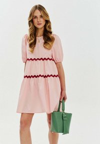 Różowa sukienka z bufiastymi rękawami i czerwonymi falbanami, z warstwowym krojem. Zielona torebka trzymana w dłoni. Gładka tekstura, swobodny krój.