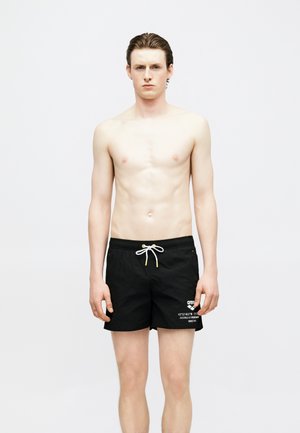 Młody mężczyzna z mokrymi włosami stoi, ubrany w czarne kąpielówki z białym sznurkiem i białym napisem na lewej nodze, na jednolitym jasnym tle.