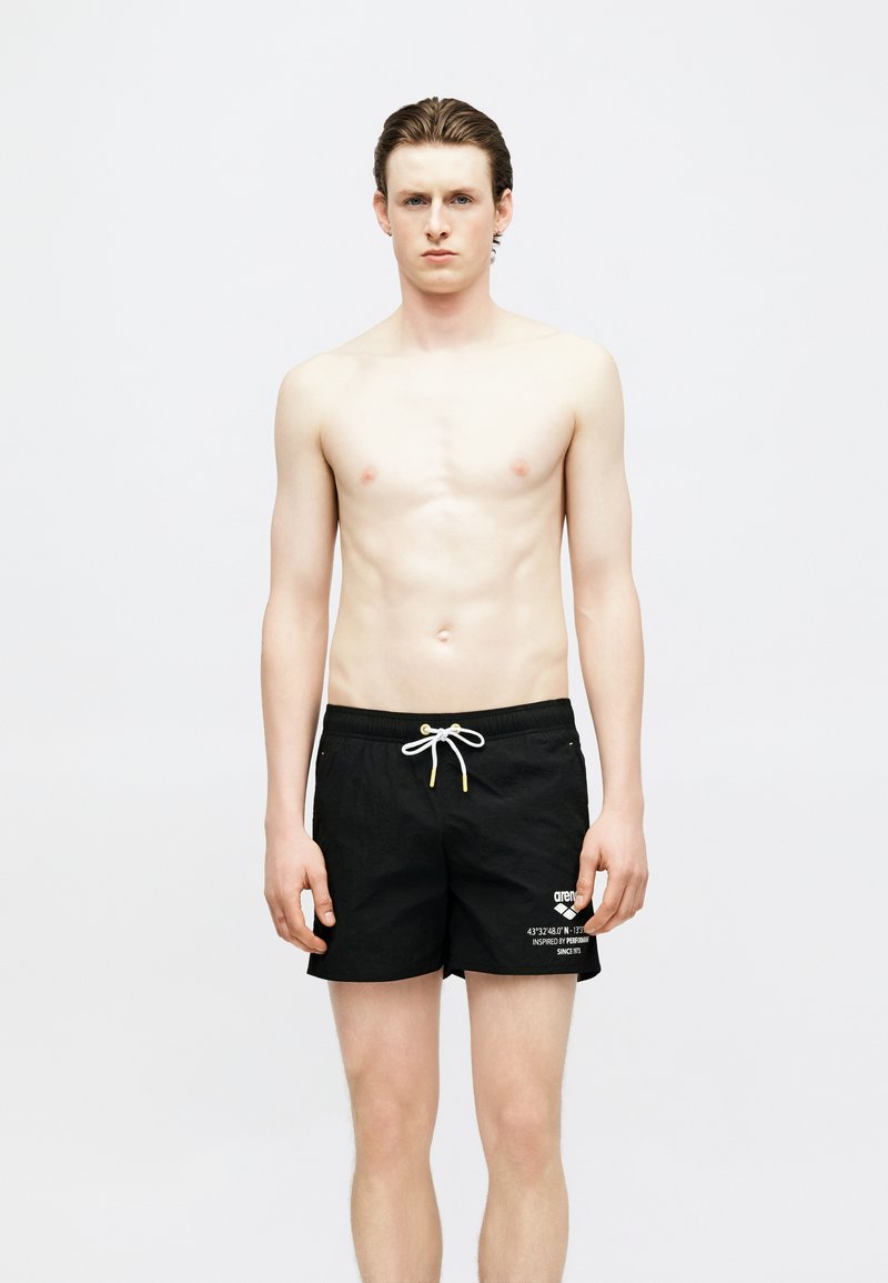 Νεαρός άνδρας με βρεγμένα μαλλιά, όρθιος, φοράει μαύρο μαγιό με λευκό κορδόνι και λευκή γραφή στο αριστερό πόδι, απλό φωτεινό φόντο.