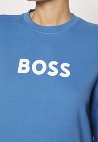 Modrá mikina z měkké látky s kulatým výstřihem a výrazným bílým logem "BOSS" na přední straně. Hladká textura a neformální design.
