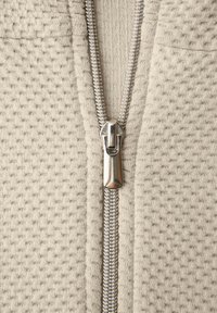 Primer plano de una cremallera de metal plateado parcialmente cerrada sobre una tela de punto texturizada beige.