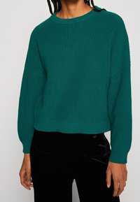 Morski dzianinowy sweter o prążkowanej fakturze, z okrągłym dekoltem i opuszczanymi ramionami, zestawiony z czarnymi aksamitnymi spodniami.