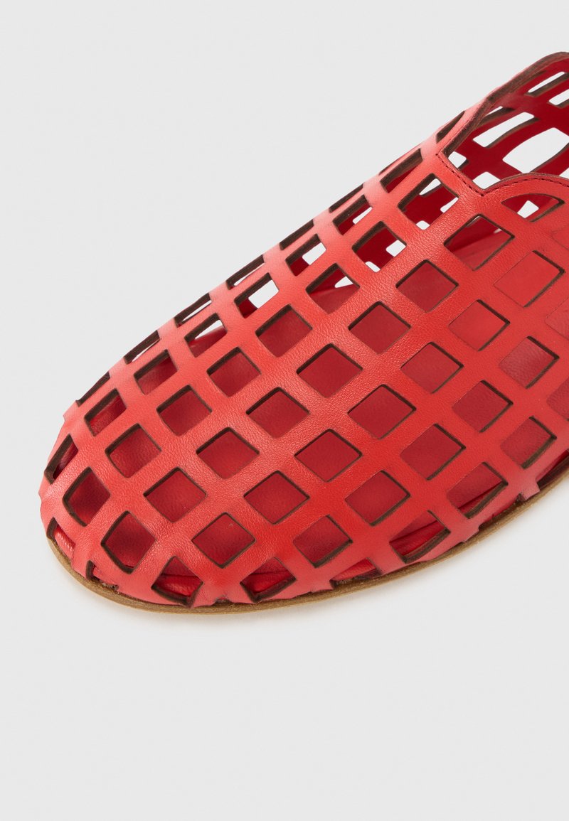 Zapato perforado rojo hecho de material flexible, con un patrón de recortes cuadrados y punta redondeada, y un forro interior suave.