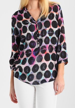 Femme portant un chemisier à manches longues avec de gros pois noirs et un fond aquarelle coloré, associé à un pantalon blanc.