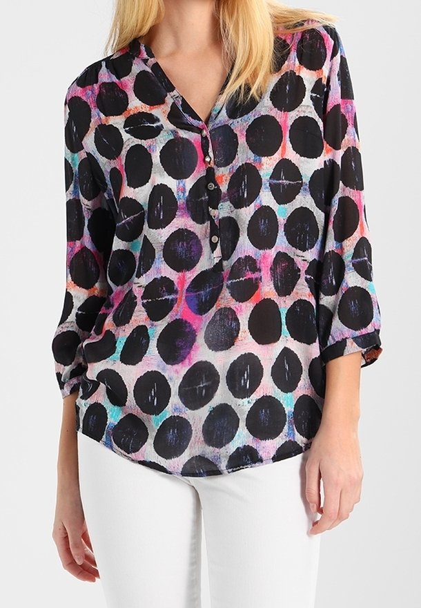 Femme portant un chemisier à manches longues avec de gros pois noirs et un fond aquarelle coloré, associé à un pantalon blanc.