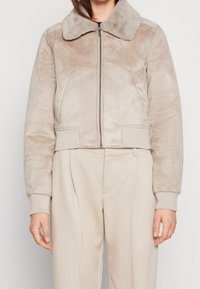 Krótką beżową kurtkę z futerkiem z imitacji o miękkiej fakturze, kontrastującym kołnierzem, zamkiem błyskawicznym i ściągaczami na mankietach, zestawioną z jasnymi spodniami.