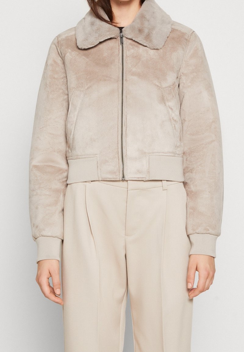 Krótką beżową kurtkę z futerkiem z imitacji o miękkiej fakturze, kontrastującym kołnierzem, zamkiem błyskawicznym i ściągaczami na mankietach, zestawioną z jasnymi spodniami.