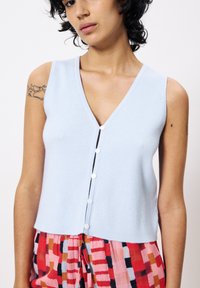 Helles, blaues, geripptes Oberteil mit V-Ausschnitt und fünf kontrastierenden weißen Knöpfen auf der Vorderseite. Weiche, gestrickte Textur.