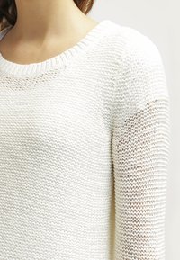 Pull en maille blanche avec un col rond et un motif texturé. Il présente un tissage lâche, laissant entrevoir la peau en dessous. Design simple et léger.
