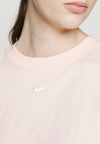 Světle růžové tričko Nike s kulatým výstřihem a bílým vyšitým logem na hrudi, vyrobené z jemné, hladké látky.