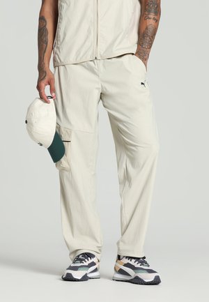 Light beige athletic pants with a relaxed fit, featuring pockets and a small black logo. Paired with multi-colored sneakers and a cream cap.