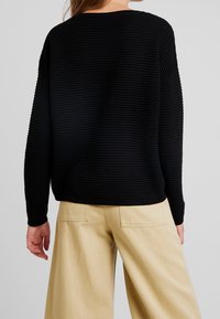 Svart ribbat stickat tröja med avslappnad passform, bred halsringning och långa ärmar, i kombination med ljusbeiga vida byxor.