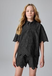 Sort broderet to-delt sæt bestående af en kortærmet skjorte med knapper, blomsterprint og bølgekant, kombineret med matchende shorts.