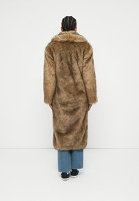 Καφέ ψεύτικο γούνινο παλτό με μεγάλο γιακά, μακριά μανίκια και χαλαρή εφαρμογή, συνδυασμένο με παντελόνι σε μπλε τζην με φαρδιές μπατζάκια και μαύρα αθλητικά παπούτσια.