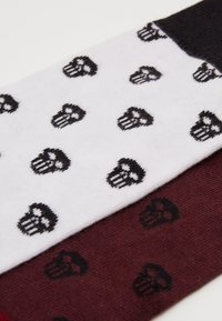Tre par strumpor: en vit med upprepat mönster av svarta dödskallar, en mörkröd med matchande dödskalle och en svart med en enfärgad design.