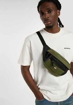 Hombre con cabello trenzado lleva una camiseta blanca de Dickies y una bolsa bandolera verde con cremallera negra y logo en la parte delantera.