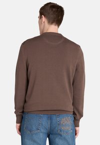 Brun stickad tröja med rund hals och ribbade ärmslut, i kombination med blåjeans som har en trädlogo broderad på bakfickan.